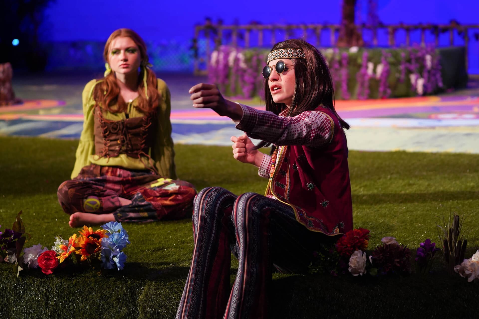 Two people in colorful, bohemian clothing sit on grass on a stage decorated with flowers. One gestures as if driving, wearing sunglasses and a headband; the other looks on, seated cross-legged, with a serious expression.