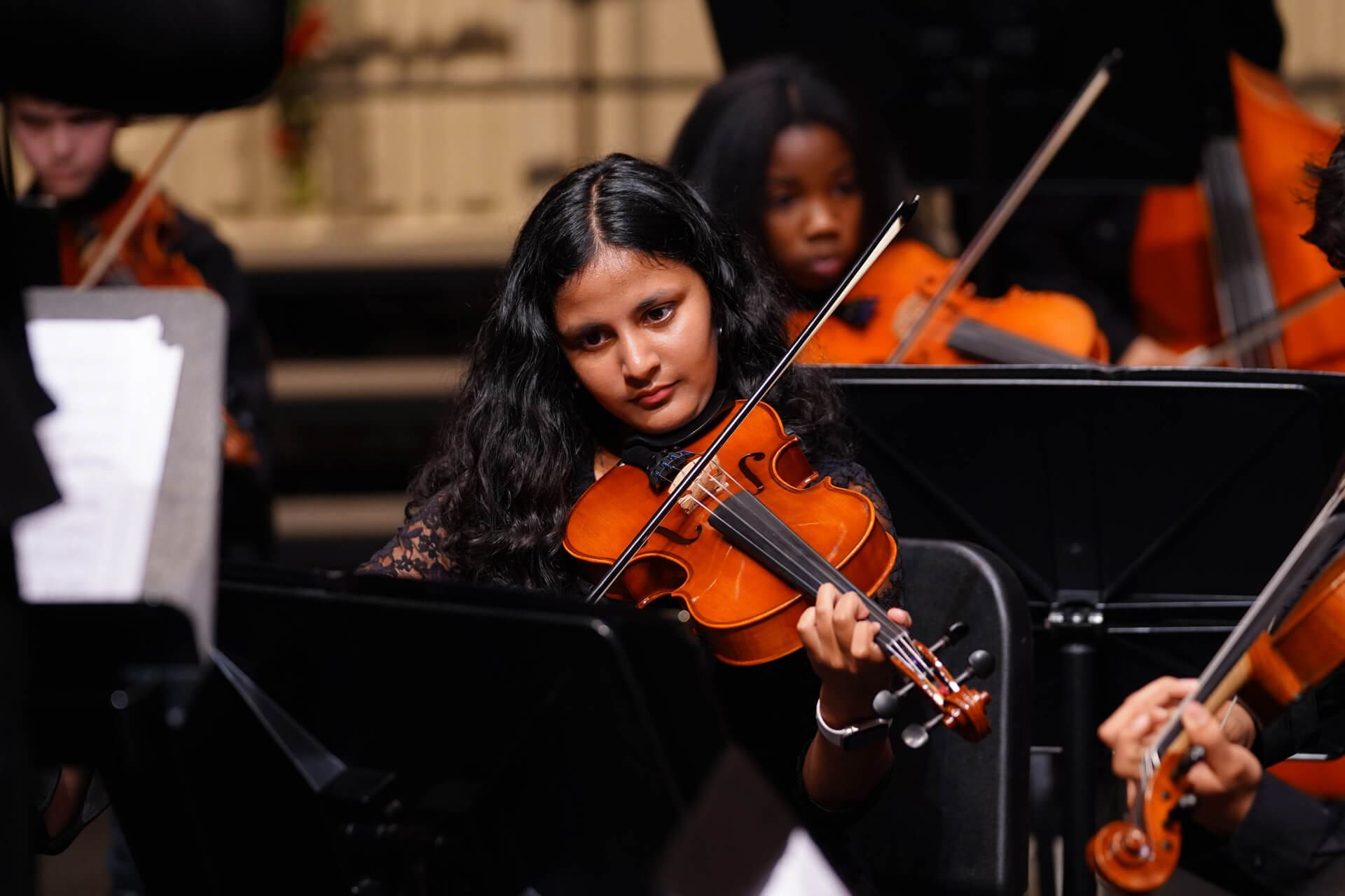 A woman with long dark hair plays the violin in an orchestra, surrounded by other musicians. She is focused on her instrument, and music stands are visible in the foreground.
