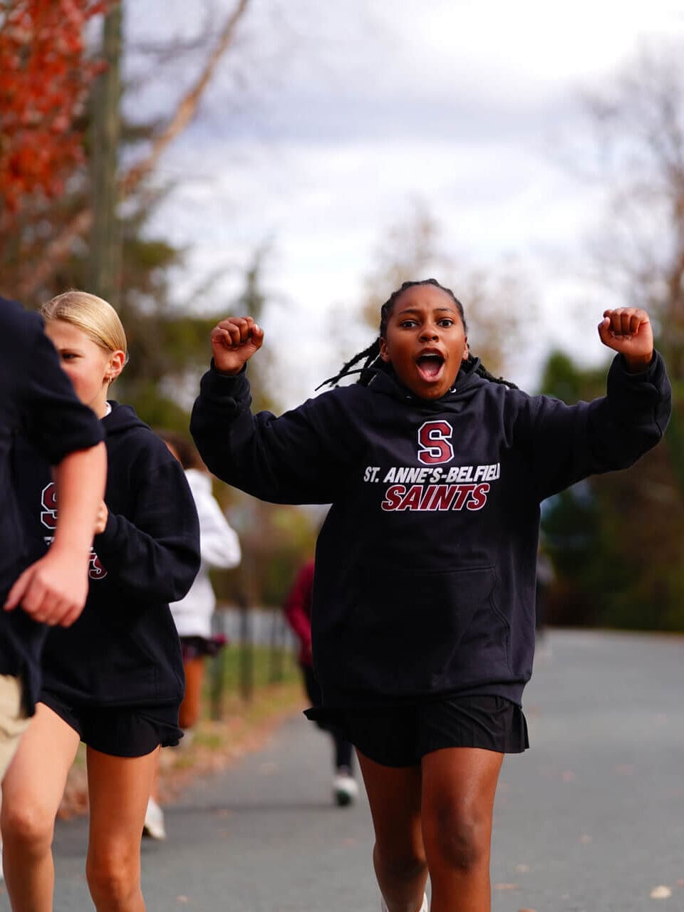 A student wearing a St. Annes-Belfield Saints hoodie raises her arms and cheers while walking outdoors with other students on a paved path.