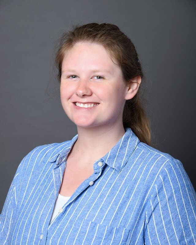 A woman with light skin and brown hair tied back smiles at the camera. She is wearing a blue and white striped button-up shirt over a white top, standing against a plain gray background.