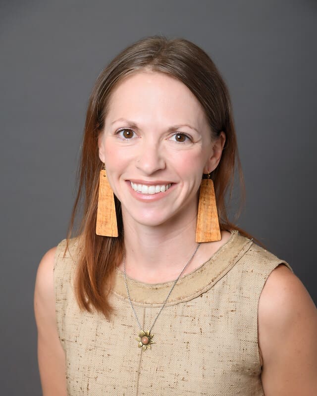 A woman with straight, light brown hair smiles at the camera. She is wearing a beige sleeveless top, large wooden rectangular earrings, and a necklace with a flower pendant. The background is plain gray.