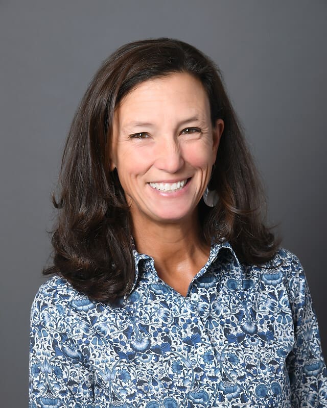 A woman with shoulder-length brown hair, wearing a blue and white patterned shirt, smiles at the camera in front of a plain gray background.