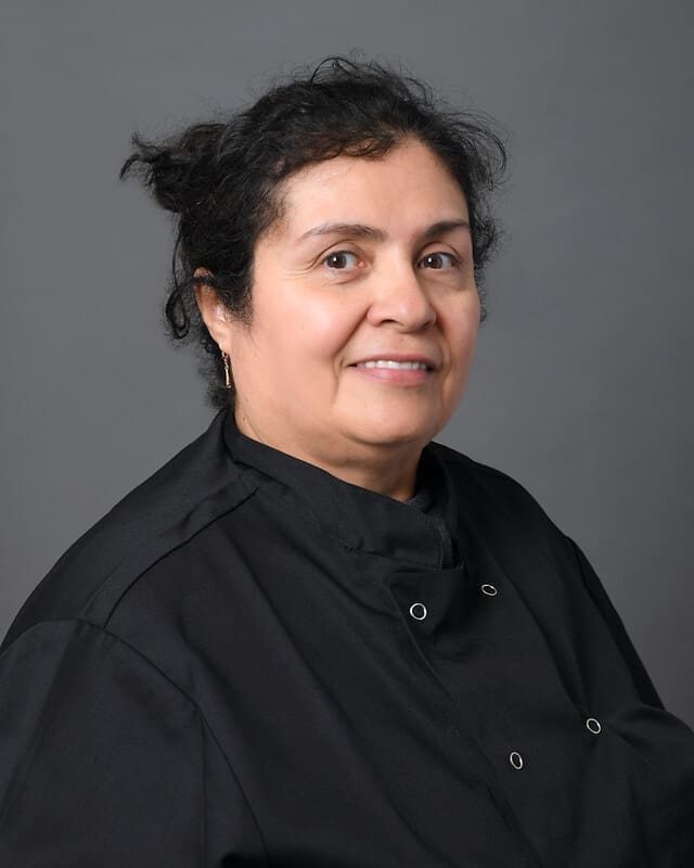 A woman with dark hair tied back, wearing a black chefs coat, poses against a plain gray background and smiles gently at the camera.