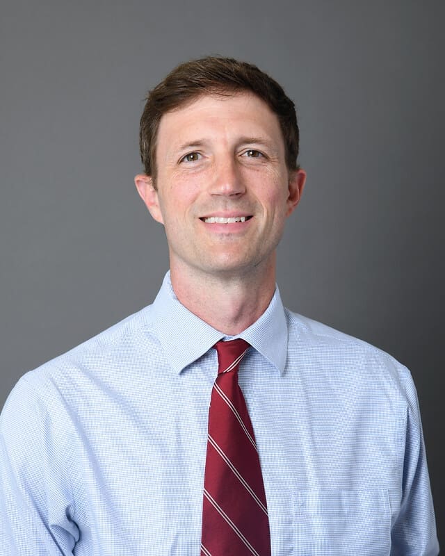 A man with short brown hair is smiling, wearing a light blue dress shirt and a red striped tie, against a plain gray background.
