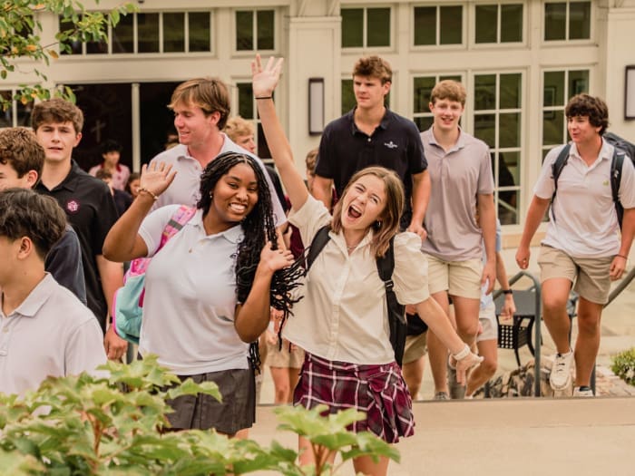 A group of high school students in uniforms walk outside a school building. Two girls in the front smile and wave enthusiastically, while others follow behind. The lively atmosphere captures a cheerful moment at school.
