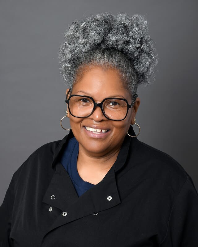 A smiling woman with gray curly hair pulled up, wearing large round glasses, gold hoop earrings, and a black buttoned top, poses in front of a gray background.