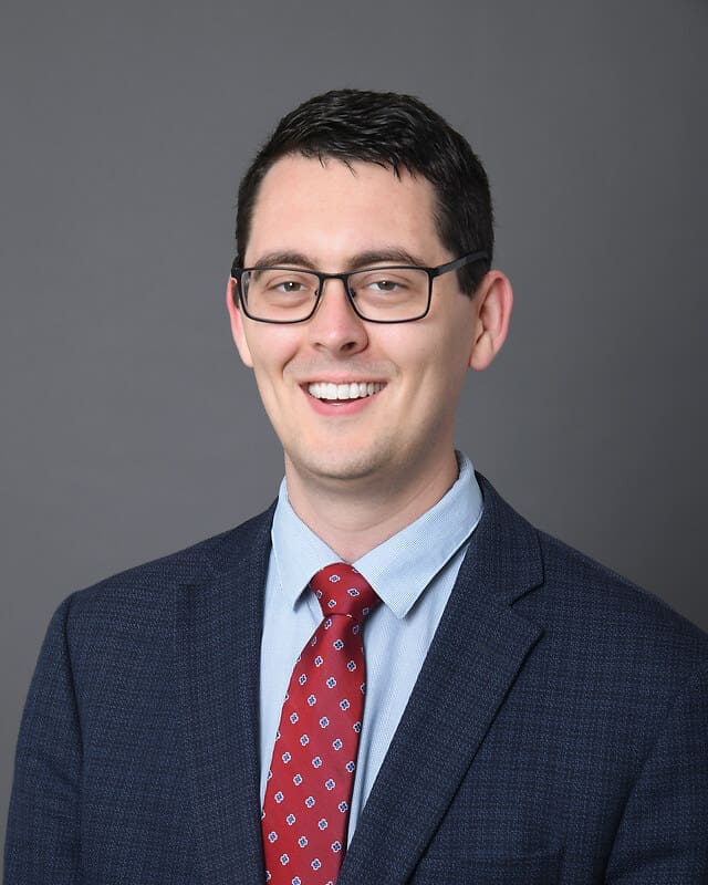 A man wearing glasses, a dark blue suit, light blue shirt, and red patterned tie smiles at the camera in front of a plain gray background.