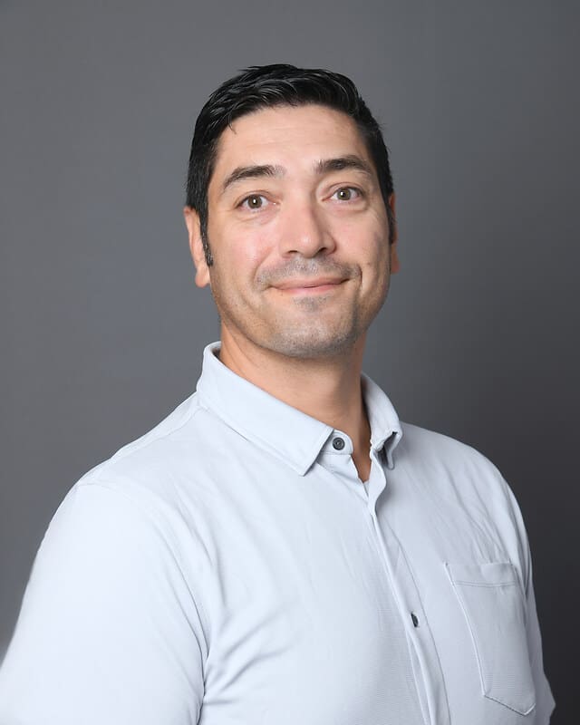 A man with short dark hair smiles slightly while wearing a light gray collared shirt against a plain gray background.