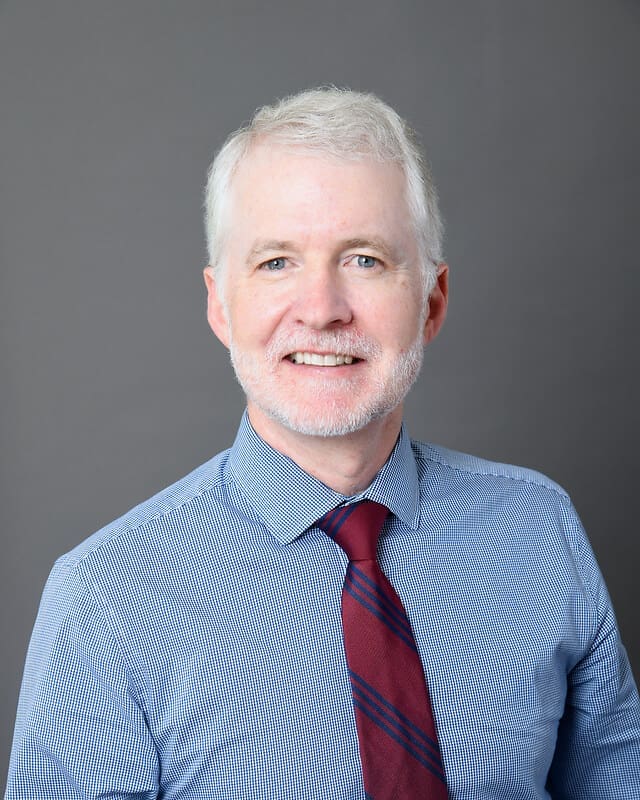 A middle-aged man with short white hair and a beard, wearing a blue checkered shirt and a red striped tie, smiles while posing against a plain gray background.