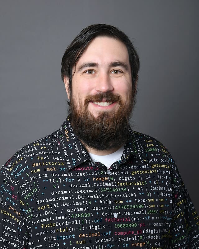 A smiling man with dark hair and a beard wears a shirt covered in colorful programming code on a gray background.