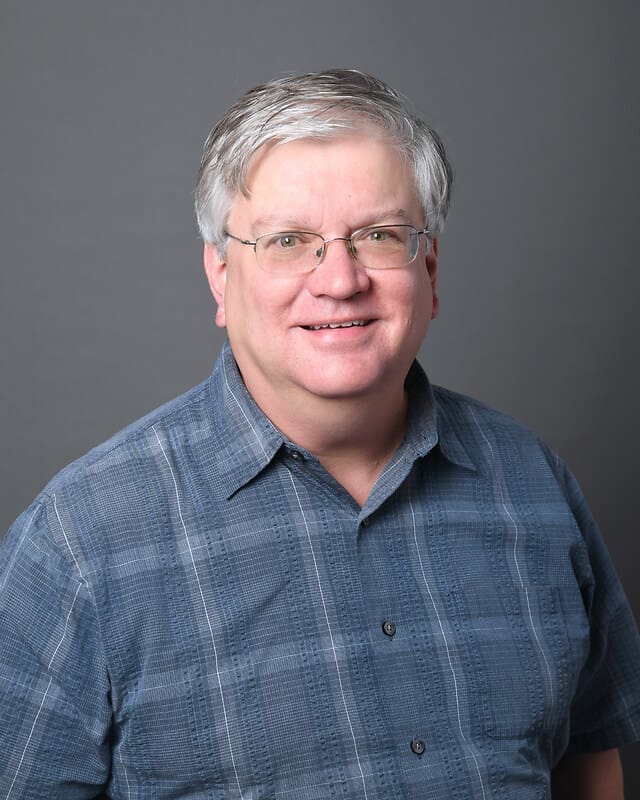 A middle-aged man with short gray hair and glasses is smiling at the camera. He is wearing a blue plaid button-up shirt and is posed against a plain gray background.