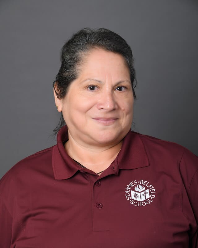 A woman with dark hair pulled back, wearing a maroon polo shirt with the St. Annes-Belfield School logo, smiles in front of a plain gray background.