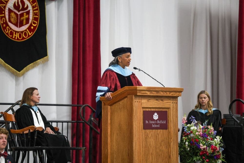 A woman in academic regalia speaks at a wooden podium during a graduation ceremony at St. Anne’s-Belfield School. Two other women in robes are seated nearby, with flowers and a school banner visible.
