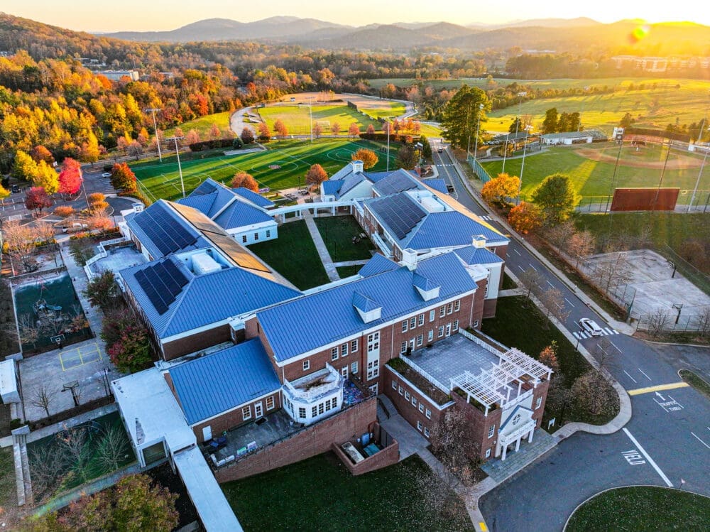 Aerial view of a large school campus with blue-roofed brick buildings, sports fields, parking lots, and surrounding trees, captured during autumn with mountains and sunrise in the background.
