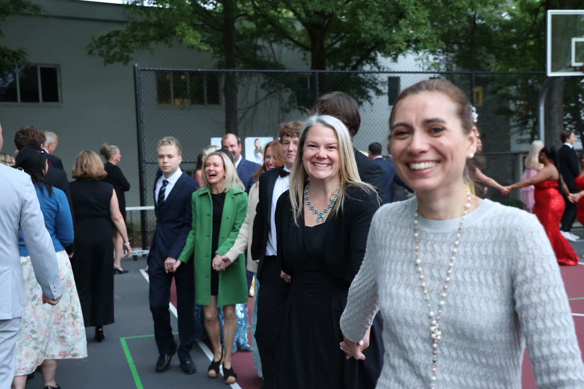 A group of people, dressed formally, smile and hold hands while walking on an outdoor school basketball court, with trees and a fence in the background.