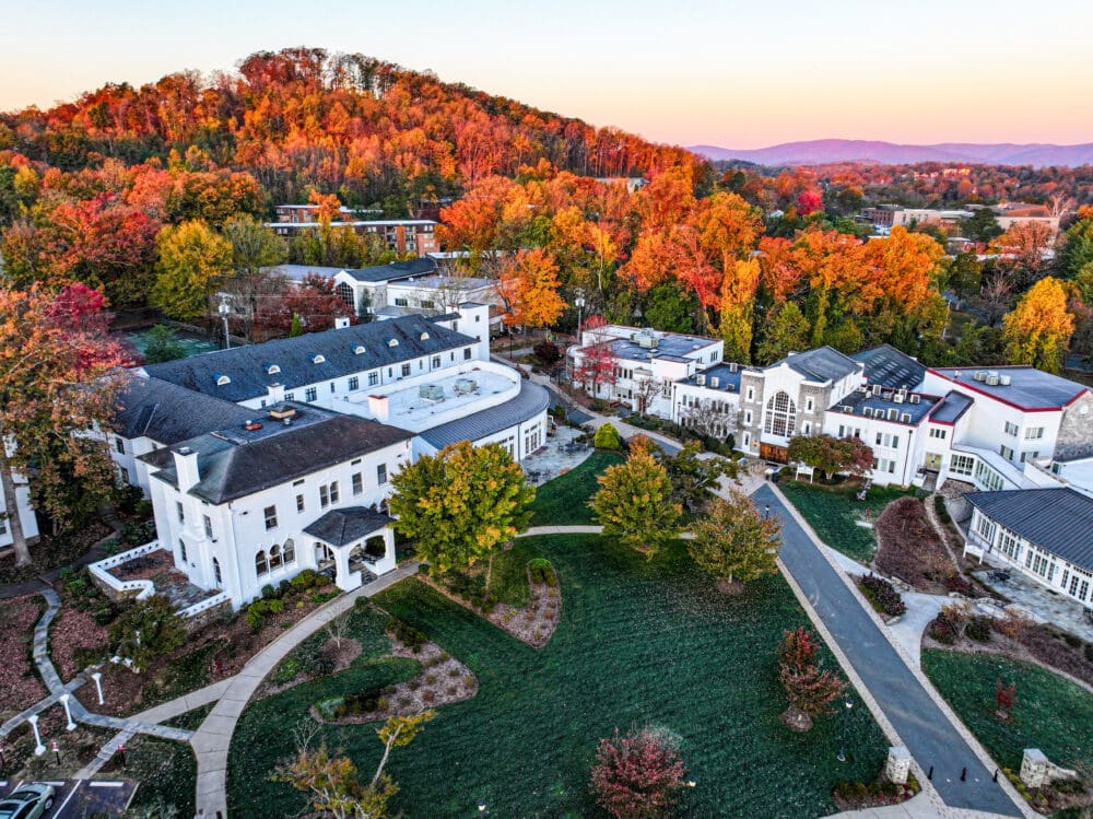 Aerial view of a campus with white buildings surrounded by vibrant autumn trees and colorful foliage, with a forested hill and mountains in the background at sunset.