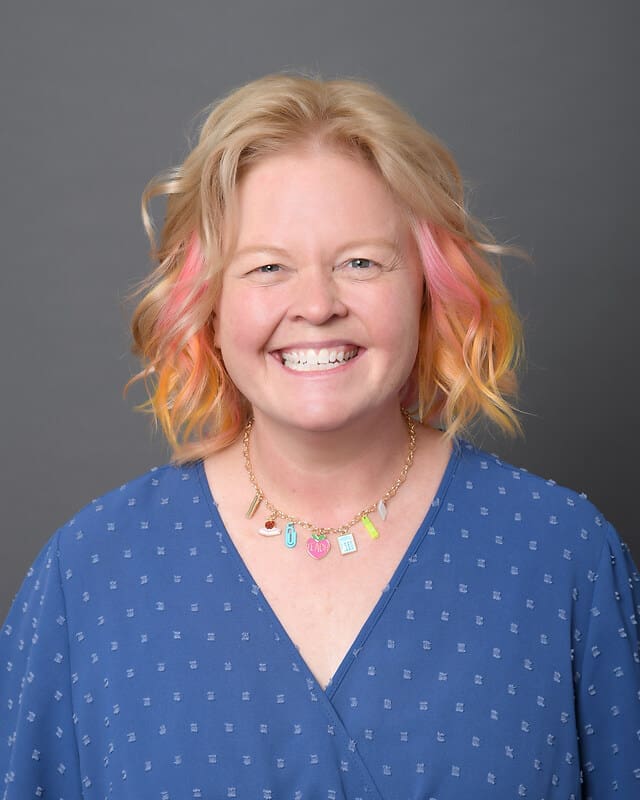 Smiling woman with wavy blonde hair with pink and orange tips, wearing a blue patterned blouse and a colorful charm necklace, standing in front of a plain gray background.