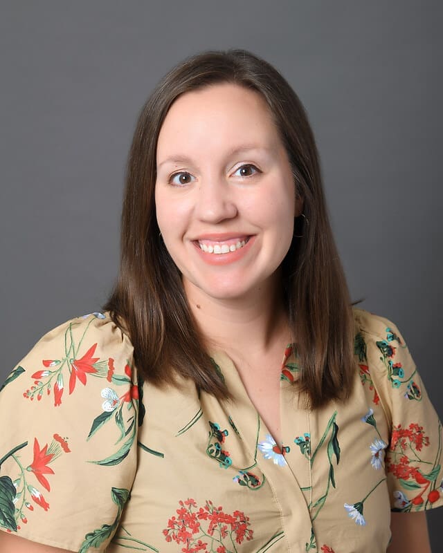A woman with straight brown hair and hoop earrings smiles at the camera. She is wearing a beige blouse with a colorful floral pattern and sits in front of a plain gray background.
