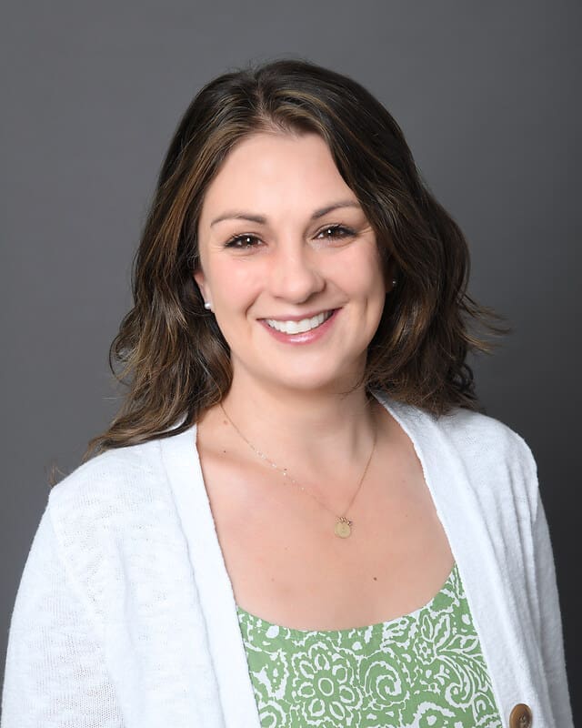 Smiling woman with wavy brown hair wearing a white cardigan over a green patterned top, posing against a plain gray background.