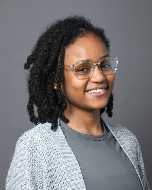 A young woman with short dreadlocks, wearing clear eyeglasses, a gray knit cardigan, and a gray shirt, smiles at the camera against a plain gray background.