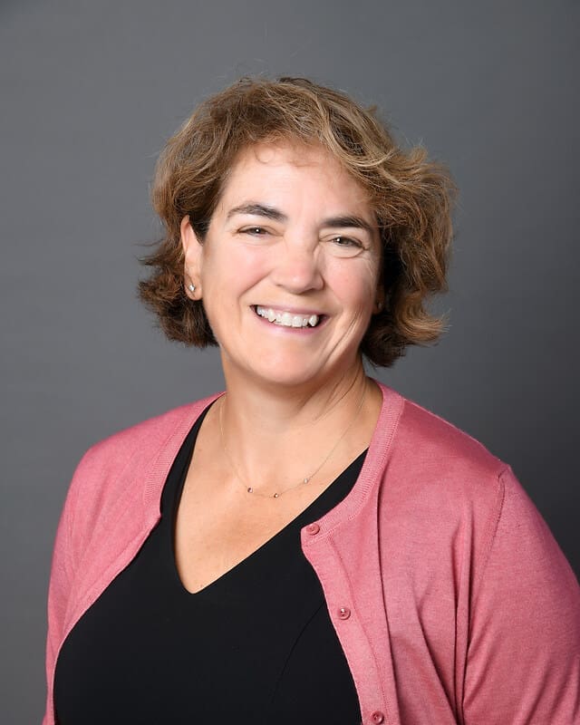 A woman with short, wavy light brown hair smiles at the camera. She is wearing a black top with a pink cardigan and a delicate necklace, posed against a plain gray background.