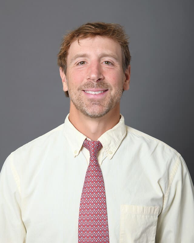 A man with short light brown hair and a trimmed beard smiles at the camera. He is wearing a cream-colored shirt and a red patterned tie, standing against a plain gray background.