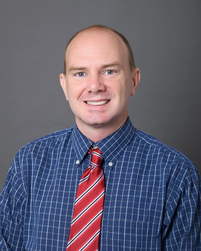 A man with short light hair is smiling at the camera. He is wearing a blue checkered shirt and a red and white striped tie. The background is plain gray.