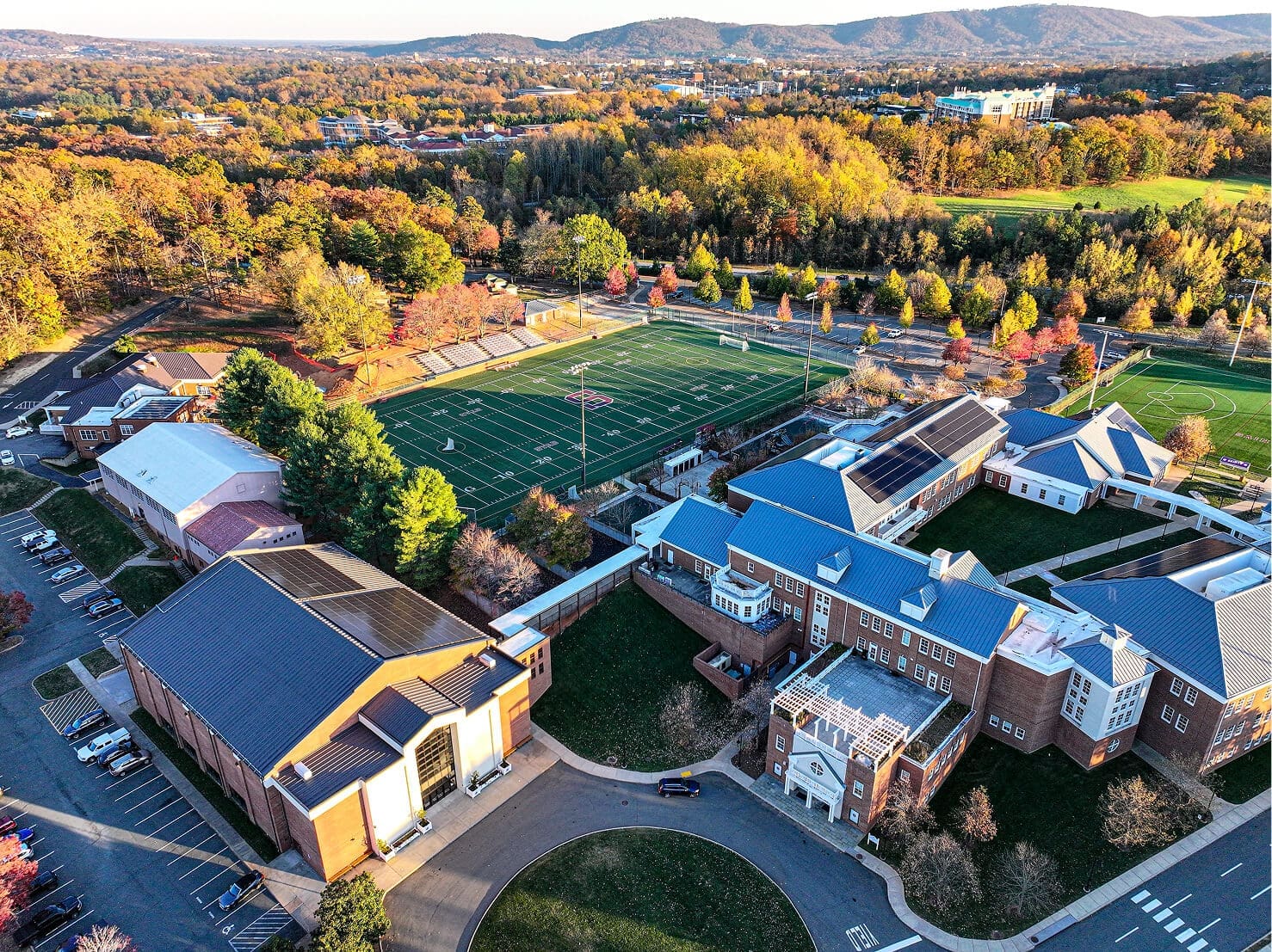 Aerial view of a school campus with brick buildings, sports fields, parking lots, and surrounding trees with fall foliage; distant hills visible in the background under a clear sky.