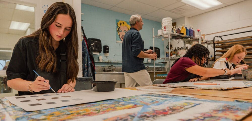 Three students work on art projects at a table in a classroom, painting and drawing, while an instructor stands in the background near shelves of art supplies.