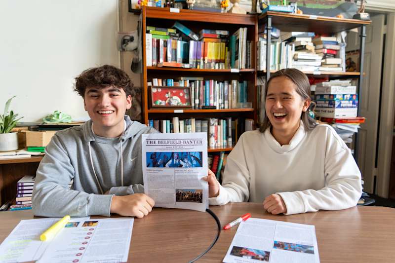 Two smiling students sit at a table with papers and highlighters, holding up a newsletter titled The Bluefield Banter in a cozy room at St. Anne’s-Belfield School, with bookshelves filled with books and decorative items in the background.