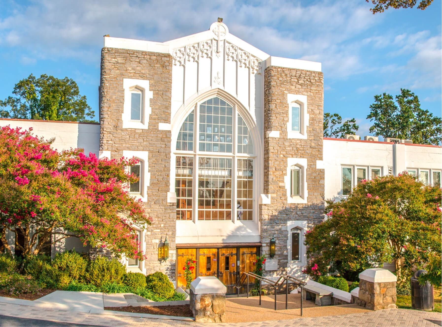 A stone and white building with large arched windows, surrounded by flowering trees and greenery, is shown on a bright, sunny day. Steps lead up to the entrance, and a blue sky with clouds is visible above.