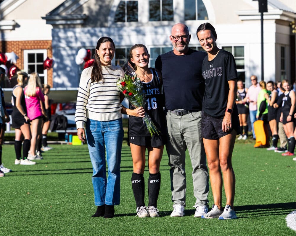 A young female field hockey player holding flowers stands with three adults on a sunny field, smiling for a photo. Other players and balloons are visible in the background.