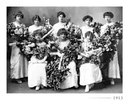 A black and white photo of eight young women in white dresses holding large bouquets of flowers, posing formally for a group portrait. The year 1913 is marked in the lower right corner.